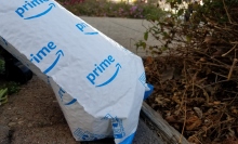 A blue and white Amazon mailer sits on the ground next to a bush. 