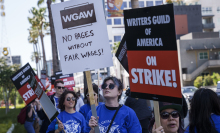 A group of people wearing blue shirts walk down a Hollywood street carrying picket signs. One sign reads "Writers Guild of America on strike!" Another sign reads "WGAW. No pages without fair wages."