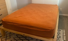 Mattress with burnt orange sheets atop a wood platform bed frame with white pillow board