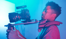 Man holding up a large video camera in a space with bright, colored lights.