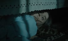A terrified looking woman hides under a bed in a dark room.