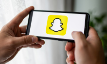 Hands holding a smart phone displaying the yellow and white Snapchat ghost logo. 