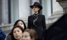 A woman in a black coat and black hat stands on steps to a courthouse.