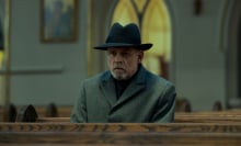 A man with dark glasses, a dark hat, and grey coat sits in a church pew.