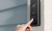 a right hand reaches toward a video doorbell mounted on the side of an external home door