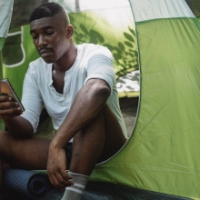 A man sits in a camping tent and looks at his phone, his backpack nearby.