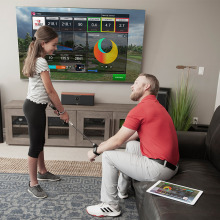 A dad sitting and watching his daughter use the TruGolf Mini Golf Simulator in front of a TV screen
