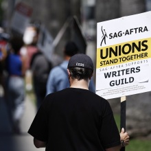 A protester in a black shirt and black baseball cap carries a picket sign that reads, "Unions stand together. SAG-AFTRA supports writers guild."