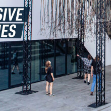 People standing underneath the 'Invasive species' installation which hangs on a metal construction outside a modern building.