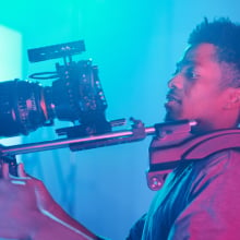 Man holding up a large video camera in a space with bright, colored lights.