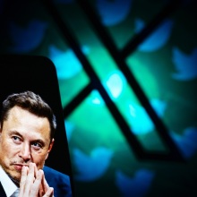 A phone screen with an image of Elon Musk held up against a background featuring the X logo.