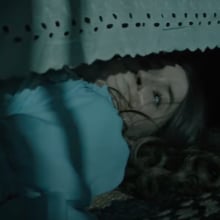 A terrified looking woman hides under a bed in a dark room.