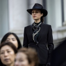 A woman in a black coat and black hat stands on steps to a courthouse.