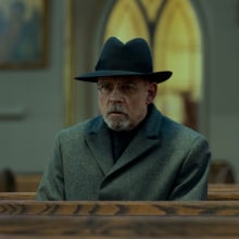 A man with dark glasses, a dark hat, and grey coat sits in a church pew.