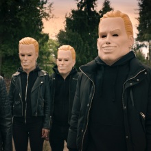 A group of people in leather jackets and rubber face masks of a smiling blonde man.
