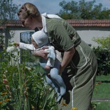 A woman holds her baby in a pretty garden, helping them smell a flower.
