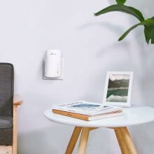 A TP Link WiFi extender is plugged into an outlet on a white wall with a side table and a chair next to it