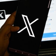 The X logo seen on a smartphone, in front of X open in a web browser on a computer screen.