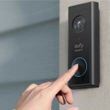 a right hand reaches toward a video doorbell mounted on the side of an external home door