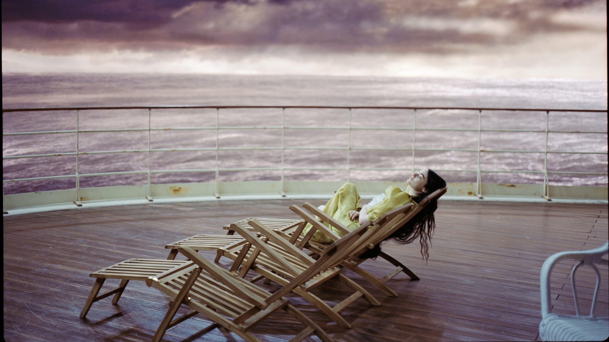 A woman with long black hair and wearing a yellow dress lounges on a chair on the deck of a ship.