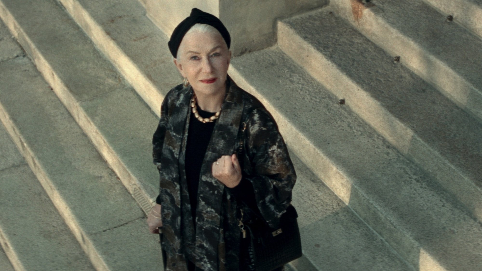 A woman in a long black coat stands outside on stone steps, smiling at the sky.