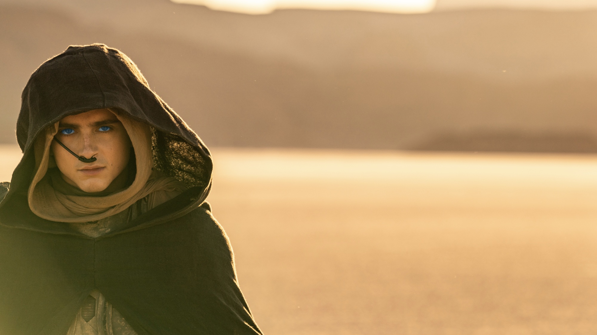 A young man with glowing blue eyes walks through the desert while wearing a brown hooded cloak.