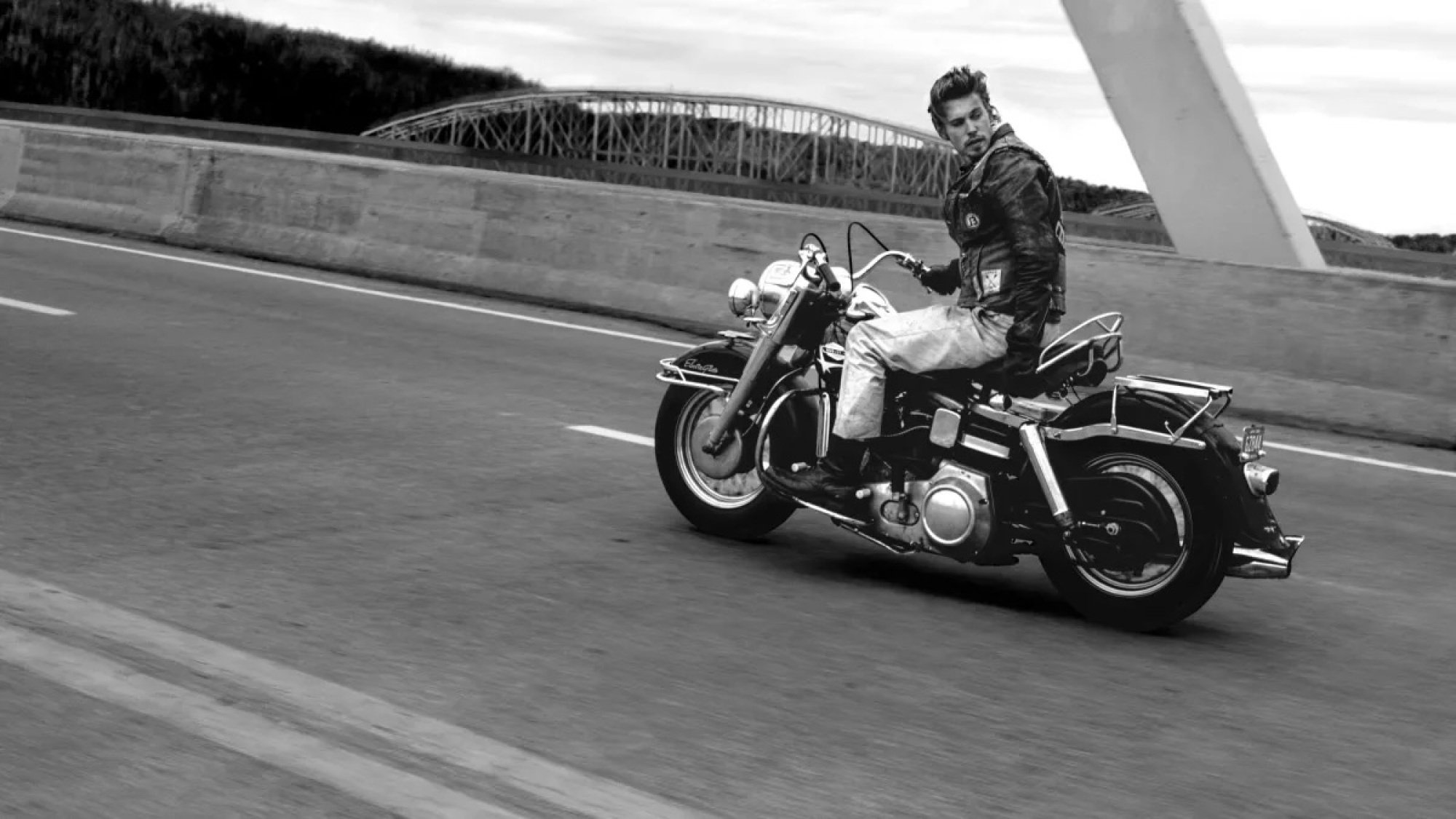 A man in a leather jacket rides a motorcycle across a bridge.