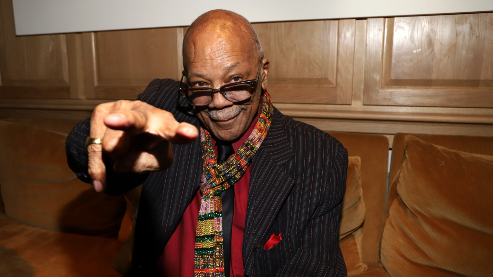 Quincy Jones sits on a leather couch pointing at the camera.