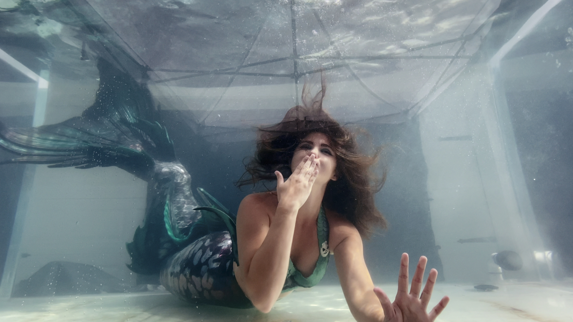 A professional mermaid blows a kiss underwater in a tank.