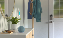 a twilight blue echo dot sitting on a white console table next to a potted plant and a wood tray filled with keys, sunglasses, and a wallet. the table is next to a door and a coat rack holding several jackets