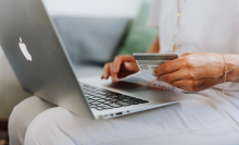 a close-up of a woman in a white top, white pants, and gold jewelry holding a credit card in front of an open macbook