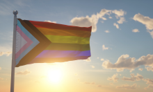 The Progress Pride flag, featuring rainbow stripes and a triangle of pink, blue, white, brown, and black stripes, waves from a flag pole over a sunny sky background