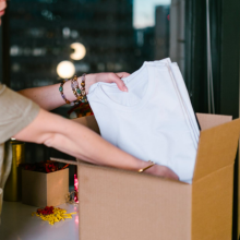 a woman in a tan polo packs a stack of white t-shirts into a cardboard box. there are four beaded bracelets on her wrist