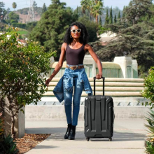 a woman in a brown tank top, jeans, and yellow sunglasses pulls a rolling samsonite suitcase through a garden on a sunny day