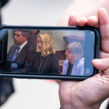A white woman's hand holding up a mobile phone as they stream the trial. Amber Heard can be see on screen.